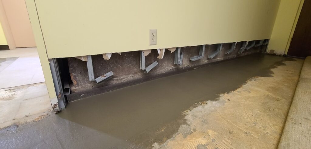 Photo of basement repair work in progress inside a building. A section of lower wall drywall has been removed, exposing metal wall supports and the concrete foundation behind it. Fresh concrete has been poured along the base of the wall where the floor meets the foundation, likely for waterproofing or structural repair. Part of the old flooring has been removed, and construction materials and exposed edges are visible around the work area.