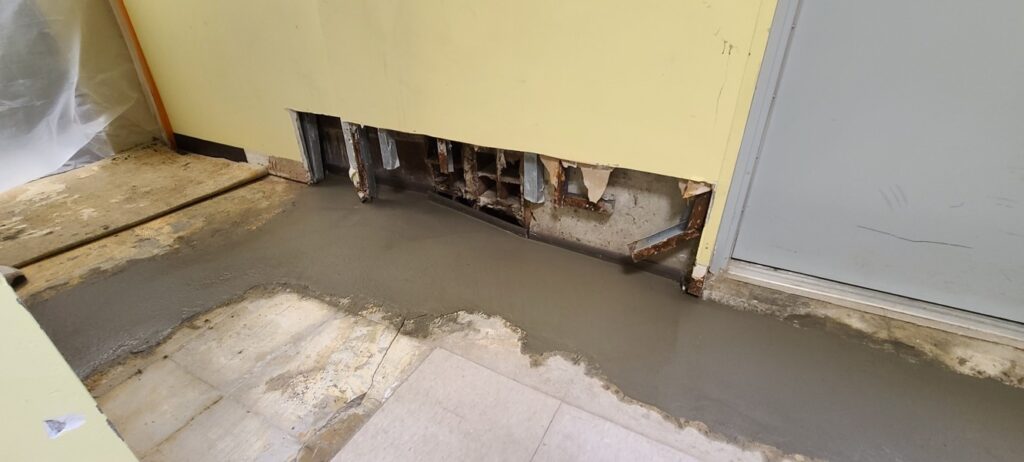 Photo of ongoing basement repair work along an interior wall. The lower portion of the wall has been cut away, exposing damaged wall materials, metal supports, and the concrete foundation behind it. Freshly poured concrete lines the edge of the floor along the wall and doorway, with sections of old flooring removed nearby. Plastic sheeting and construction debris indicate active waterproofing or foundation repair work in progress.