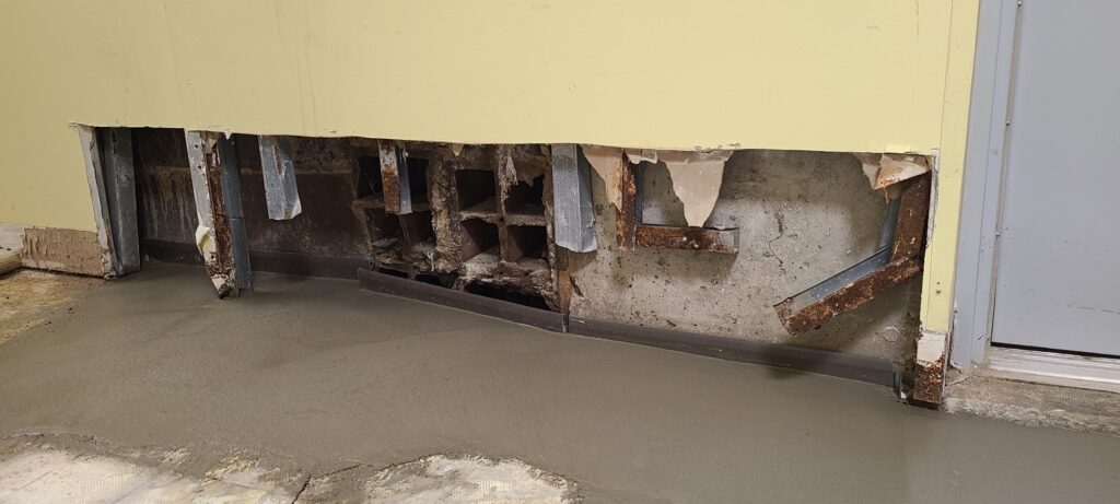 Close-up photo of basement wall repair work in progress. The lower section of the wall has been removed, exposing rusted metal framing, damaged interior materials, and sections of the concrete foundation behind it. Fresh concrete has been poured along the base of the wall where the floor meets the foundation, likely as part of a waterproofing or drainage repair project. Partially removed flooring and construction wear are visible around the repair area.